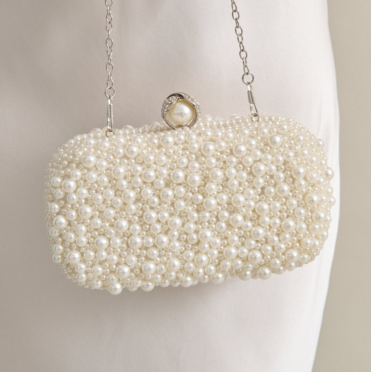 A close-up of a person in a white satin dress holding a pearl-beaded evening clutch bag with a silver chain strap and pearl clasp.