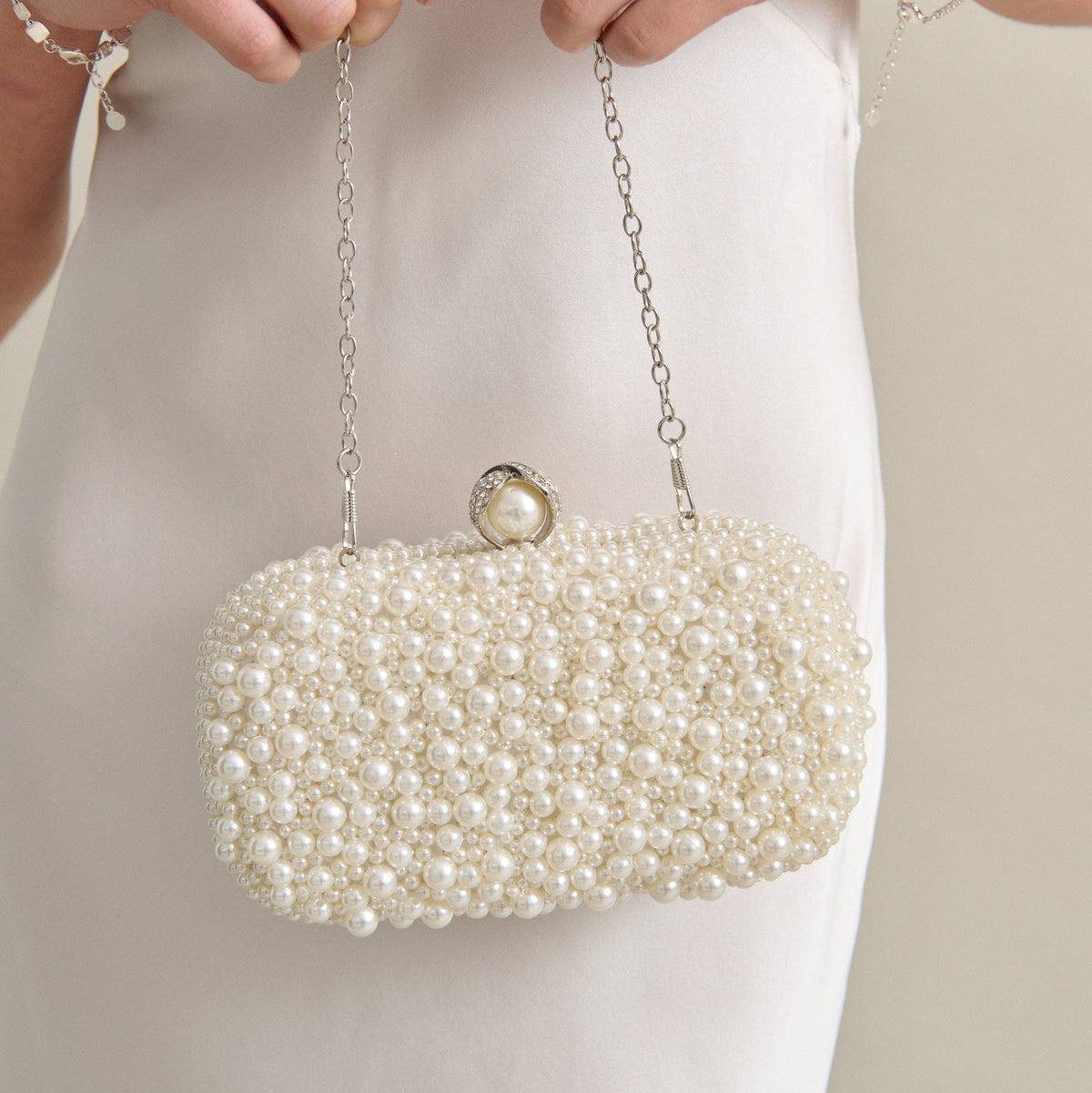 A close-up of a person in a white satin dress holding a pearl-beaded evening clutch bag with a silver chain strap and pearl clasp.